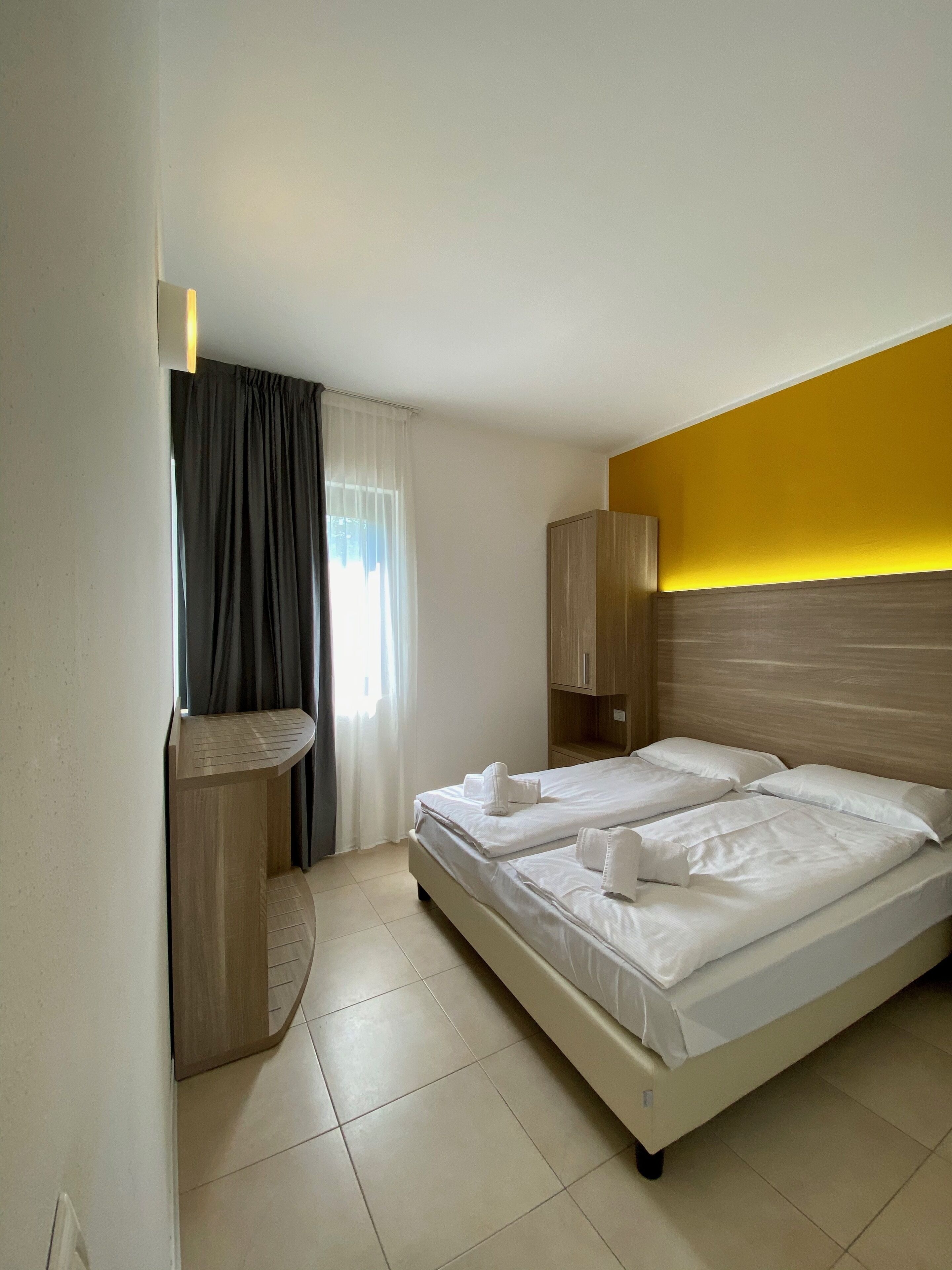 Una habitació d'hotel amb un llit, una petita taula de tocador amb mirall i una finestra amb cortines.
