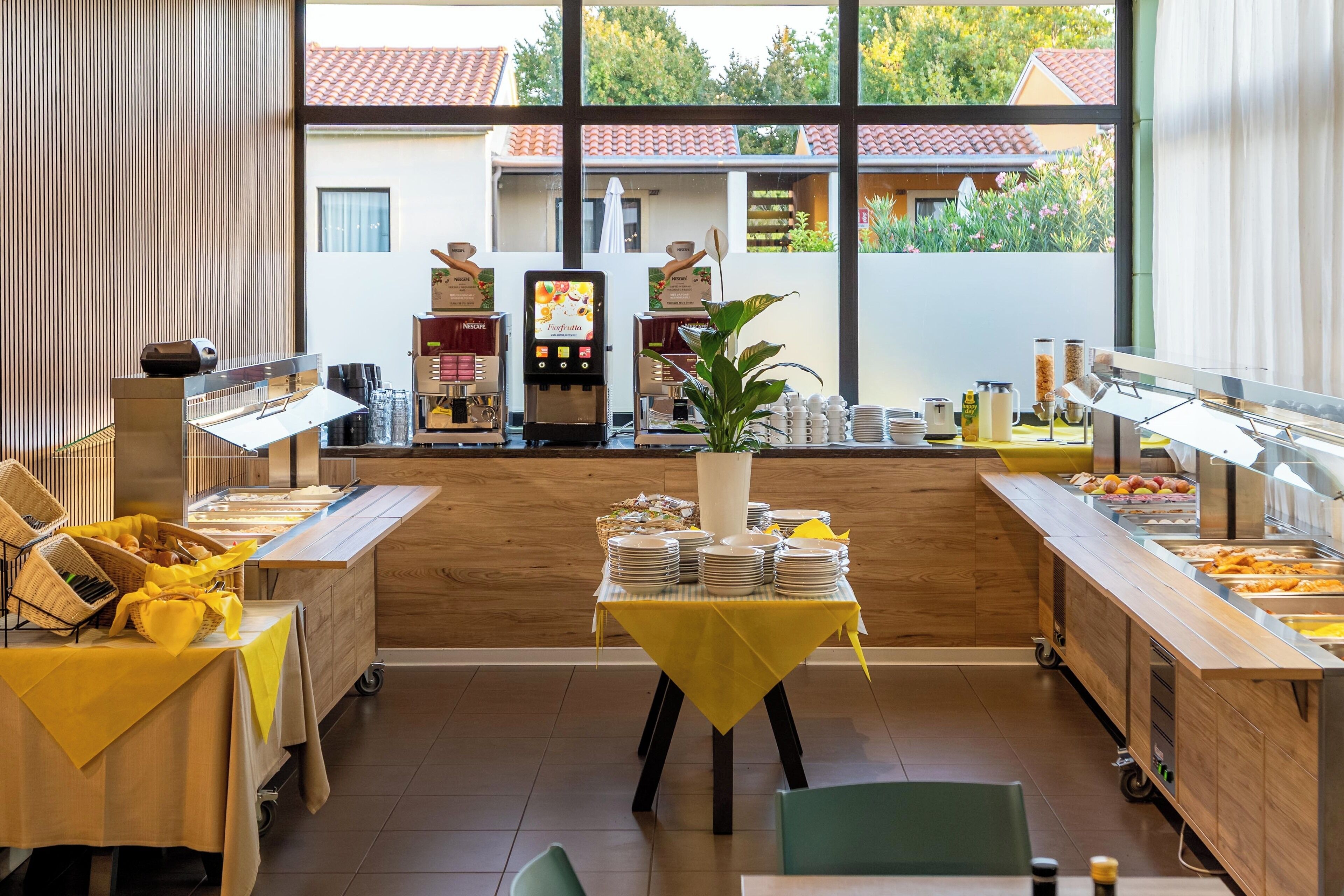 Ett område med frukostbuffé, med stationer där man själv kan plocka mat, ett centralt serveringsbord och utsikt över omgivningarna genom stora fönster.