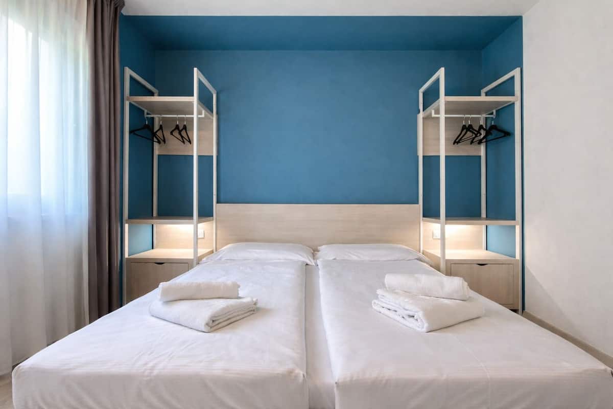 Een tweepersoonsbed met witte lakens en handdoeken, twee nachtkastjes en inbouwkasten met hangende kleding.