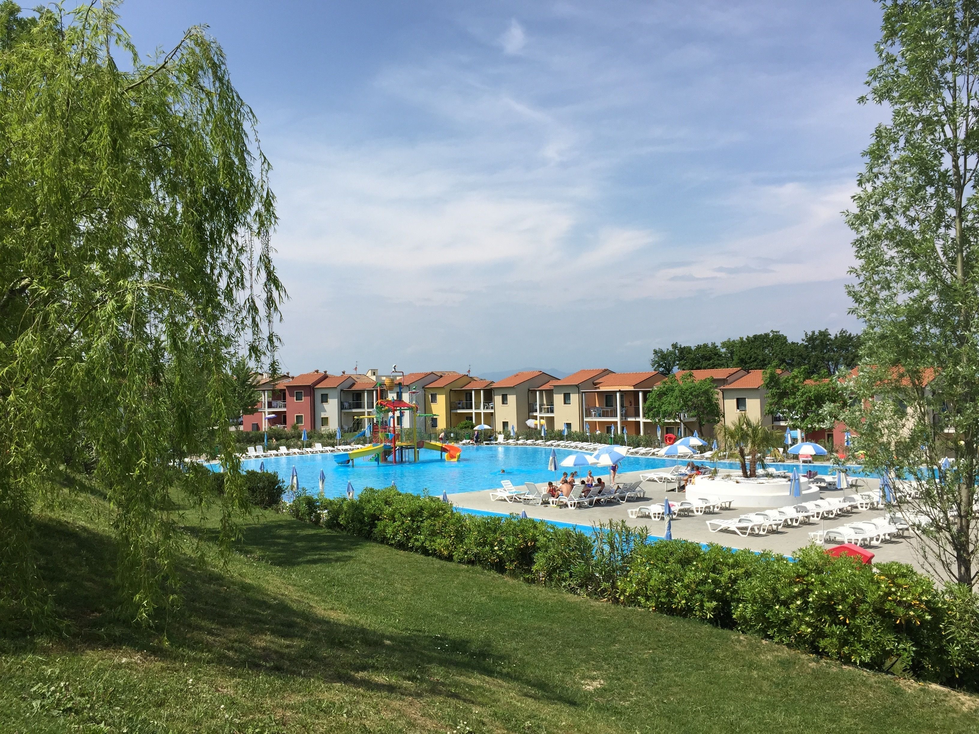 Zona de piscina con múltiples sillas de descanso y sombrillas, rodeada de edificios de varios pisos con tejados rojos.