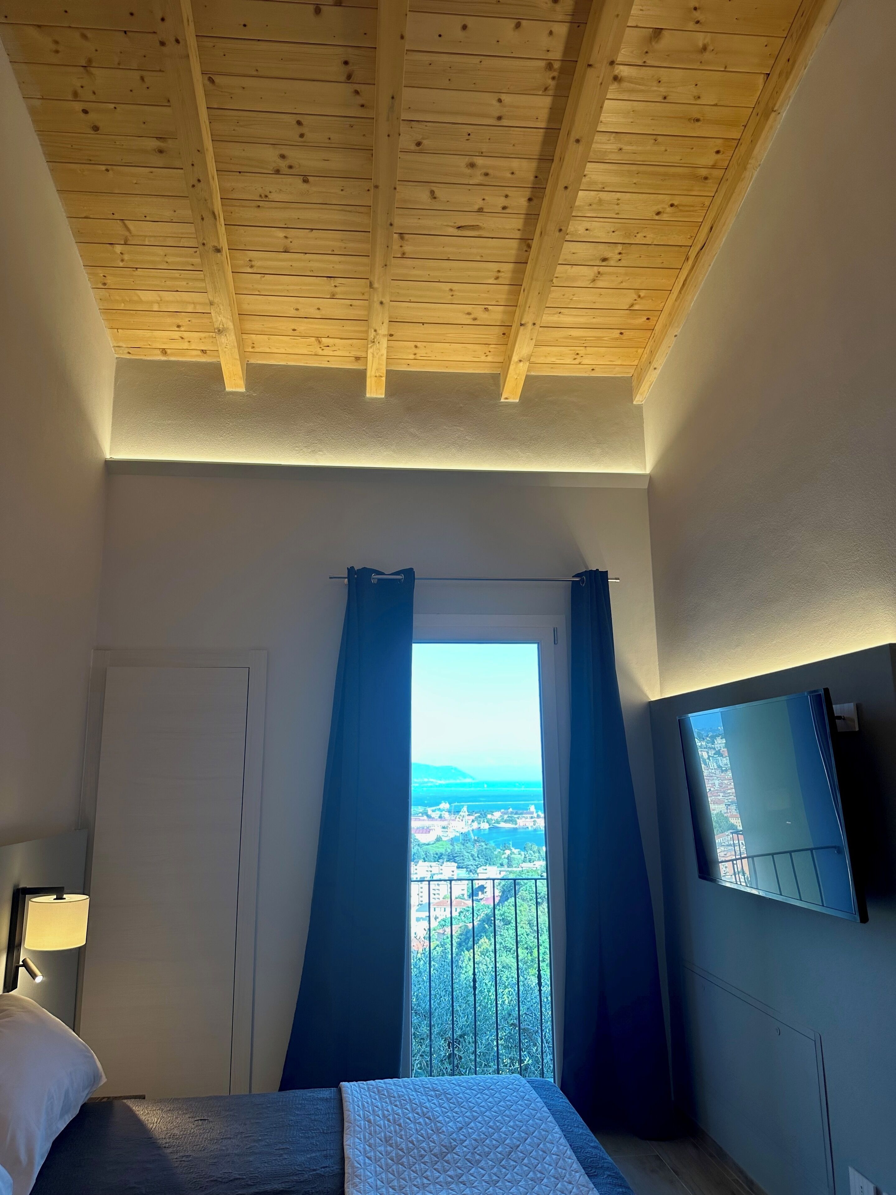 Une chambre avec un plafond en bois, un lit, un téléviseur fixé au mur et une fenêtre avec des rideaux.