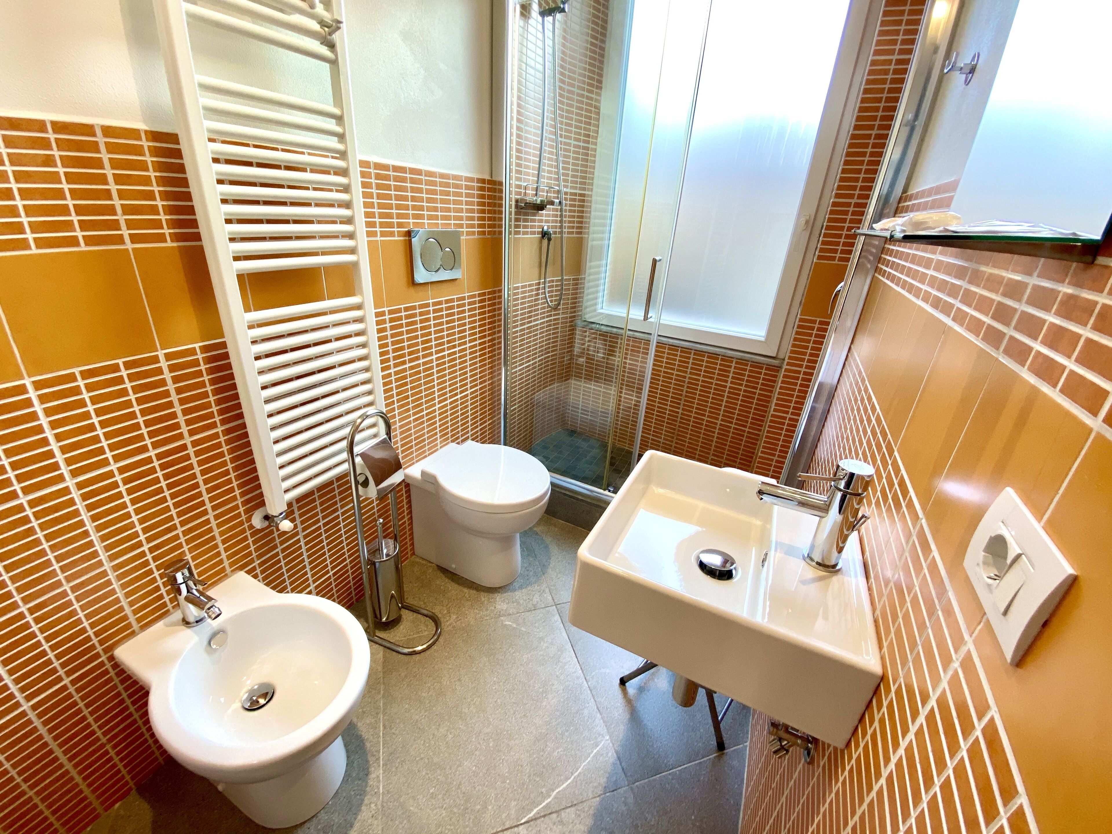 Une salle de bain équipée de toilettes, d’un bidet et d’un lavabo, avec des murs recouverts de carreaux orange et une douche à parois de verre.