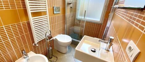 Une salle de bain équipée de toilettes, d’un bidet et d’un lavabo, avec des murs recouverts de carreaux orange et une douche à parois de verre.