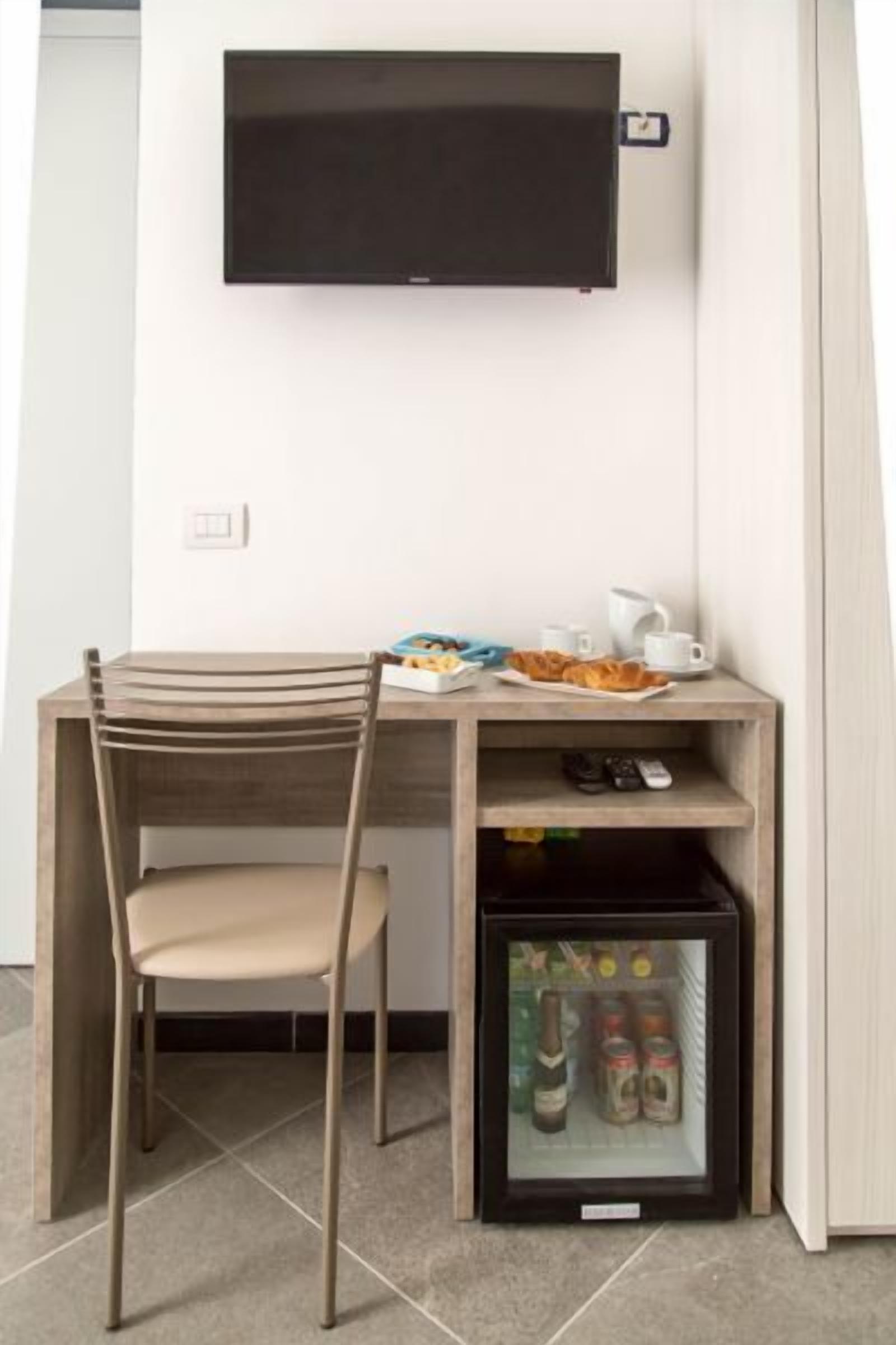 A compact room with a wooden desk, a chair, a television mounted on the wall, and a mini-fridge below the desk containing beverages.