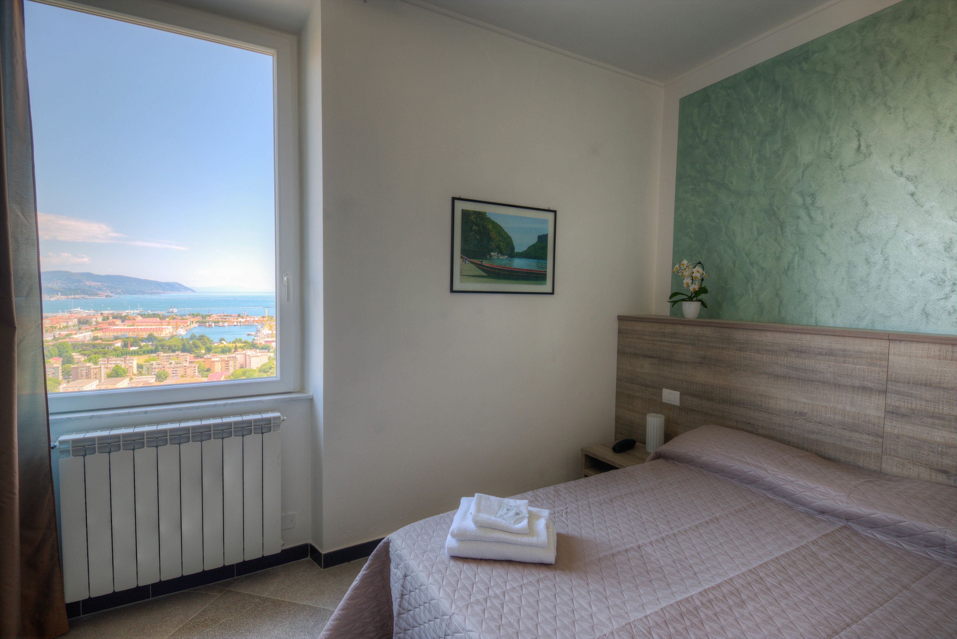 A hotel room with a bed, a view of a coastal city, a framed picture on the wall, and a radiator.