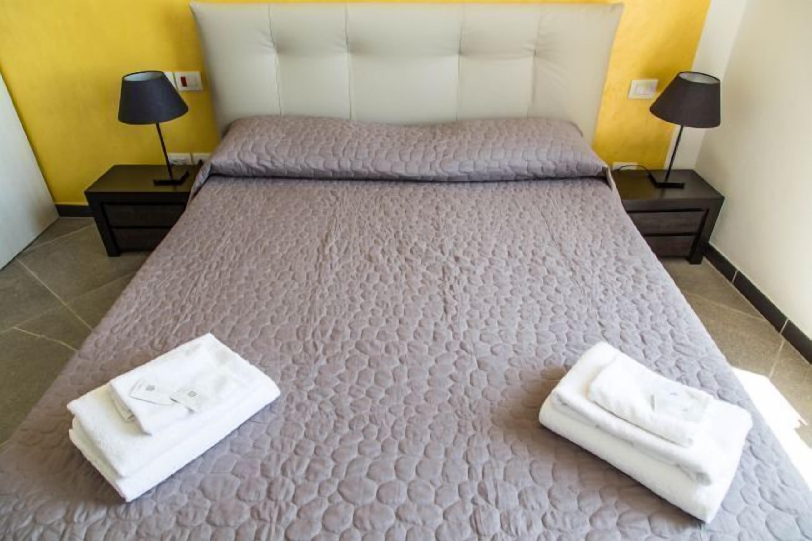 Un lit bien fait, recouvert d’une couette grise, deux tables de chevet avec des lampes et des serviettes pliées posées sur le lit.