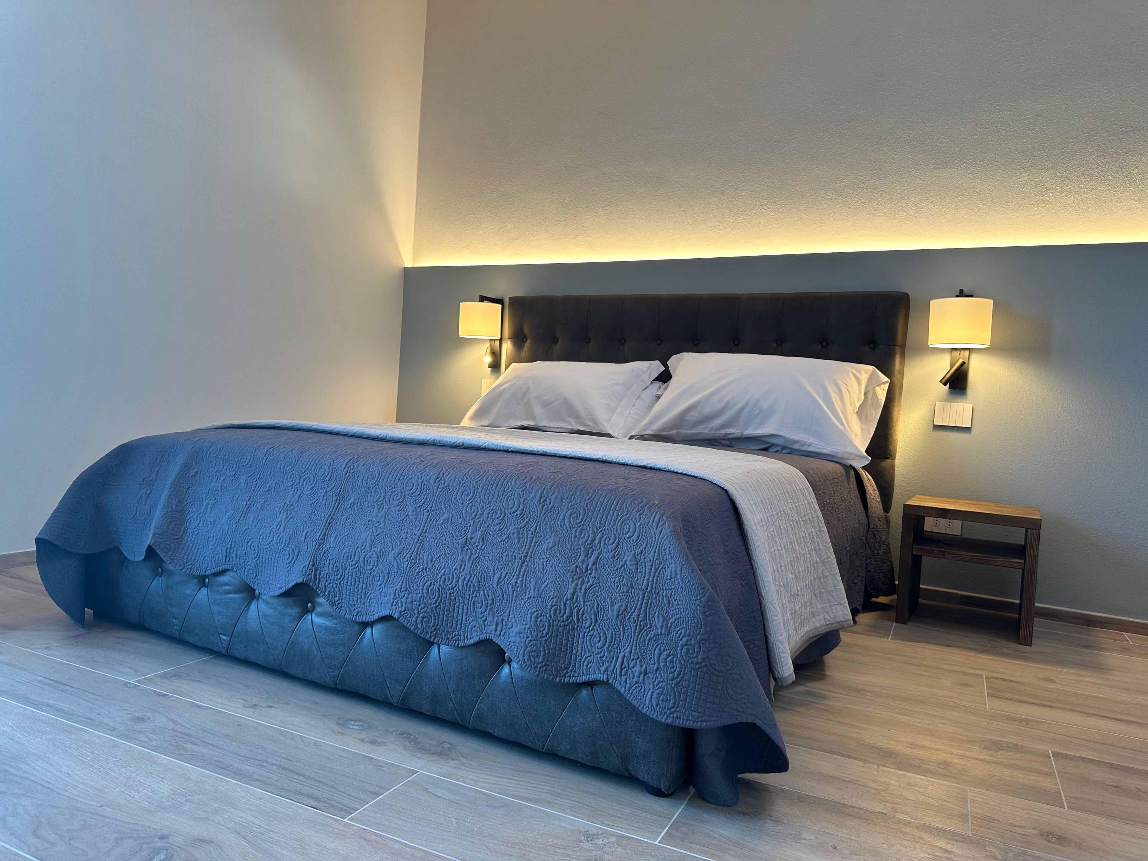 A neatly made bed with a grey upholstered headboard and a matching grey bedspread. There are two bedside lamps on either side of the bed.
