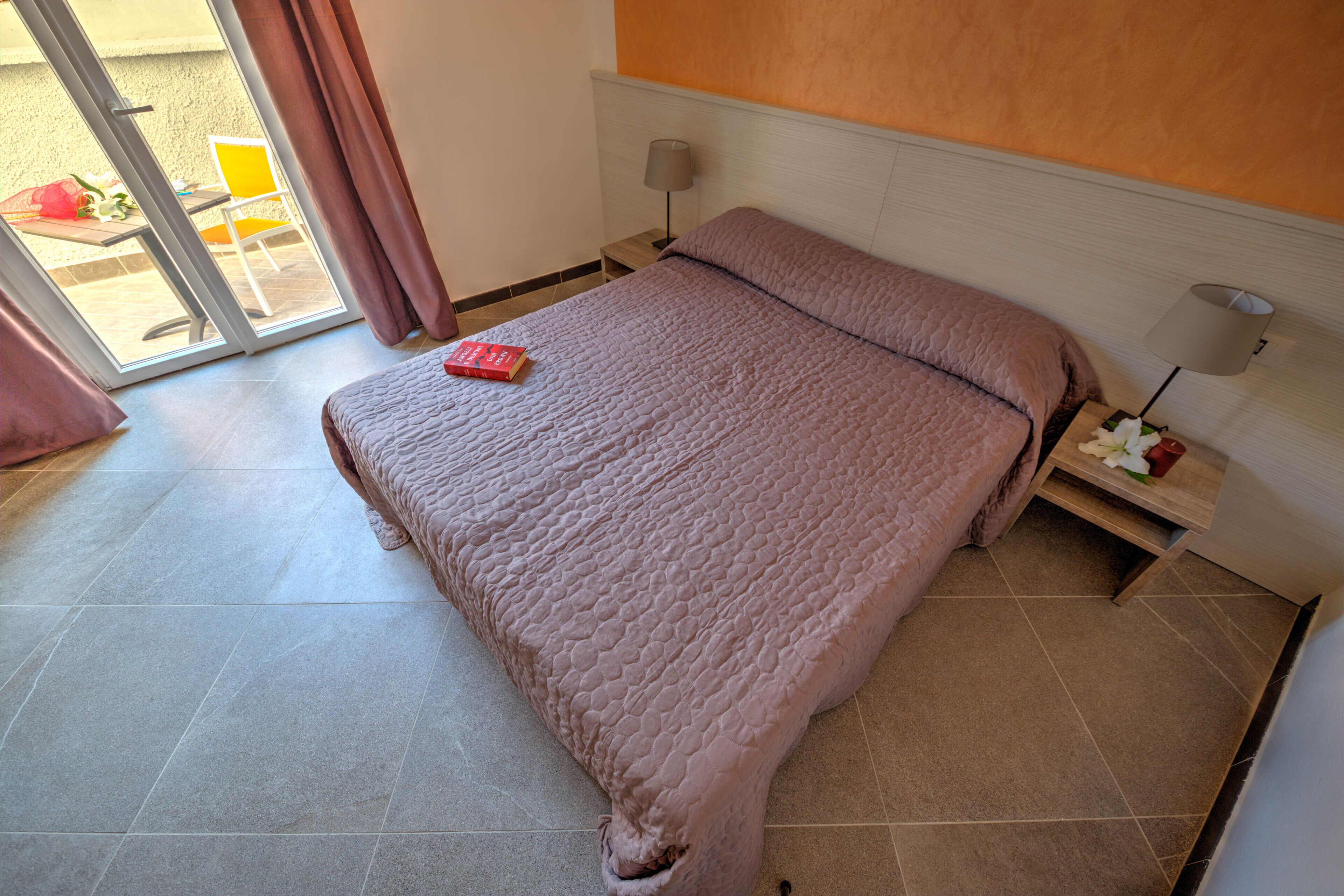 Un dormitorio con cama, mesita de noche, lámpara y vista al exterior.