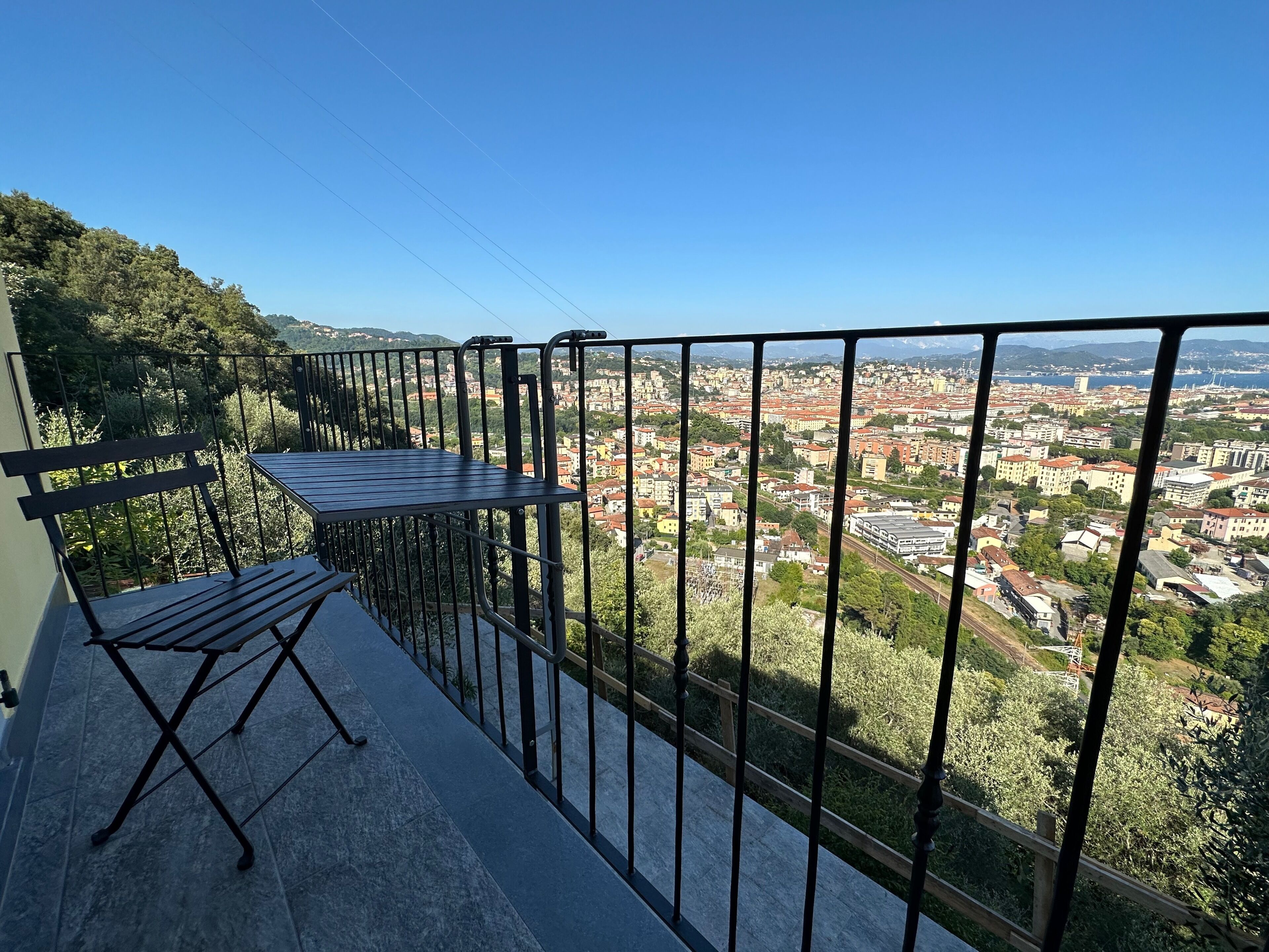 A balcony with a table and chairs, overlooking a cityscape with buildings and greenery.