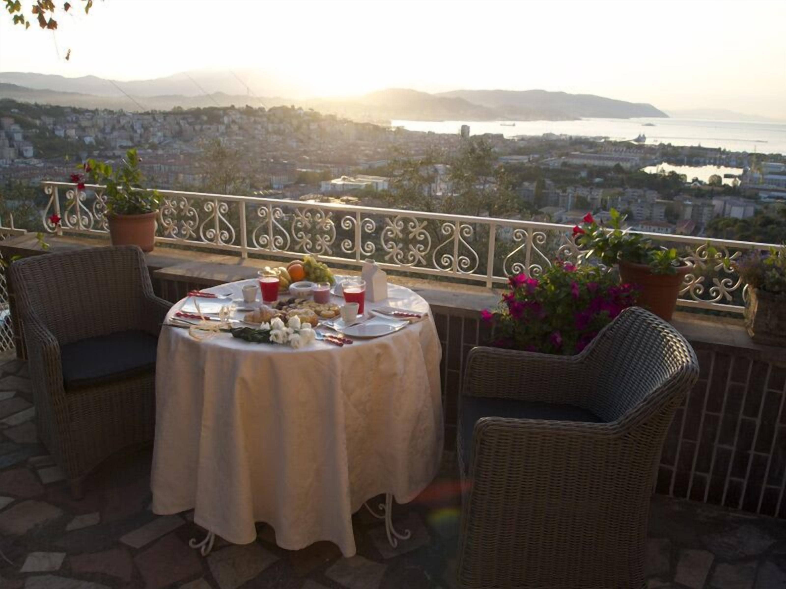 A balcony with wicker chairs, a table set for a meal, potted plants, and a cityscape view in the background.