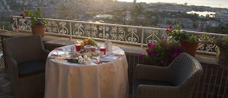 Un balcón con sillas de mimbre, una mesa preparada para una comida, plantas en macetas y una vista panorámica de la ciudad al fondo.