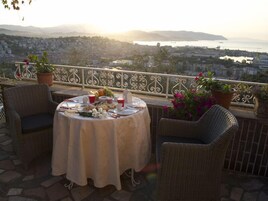 Un balcón con sillas de mimbre, una mesa preparada para una comida, plantas en macetas y una vista panorámica de la ciudad al fondo.