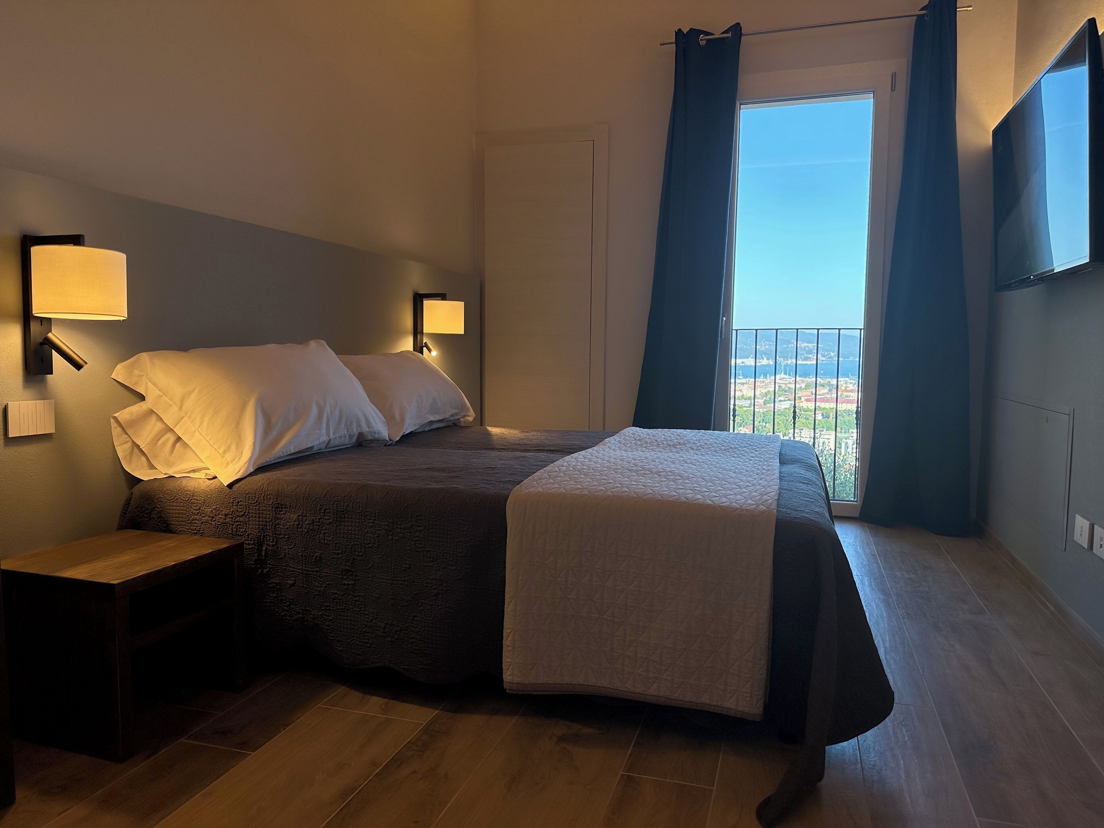 A bedroom with a bed, bedside lamps, a nightstand, a window with curtains, and a view of the outside.