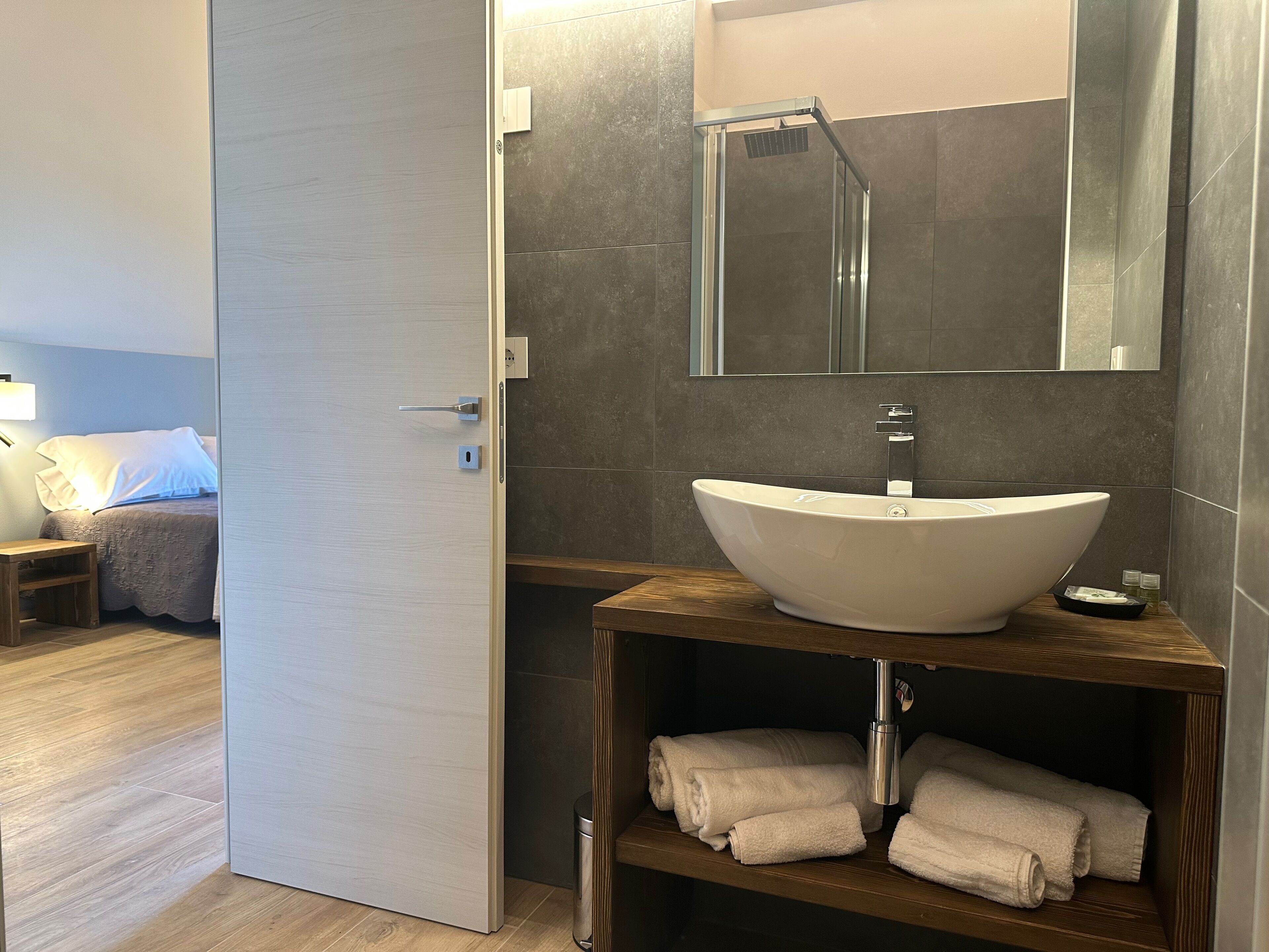 Une salle de bain moderne avec un grand lavabo sur pied, un miroir au-dessus et une cabine de douche. On y trouve une étagère en bois avec des serviettes et une petite poubelle.