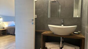 Une salle de bain moderne avec un grand lavabo sur pied, un miroir au-dessus et une cabine de douche. On y trouve une étagère en bois avec des serviettes et une petite poubelle.