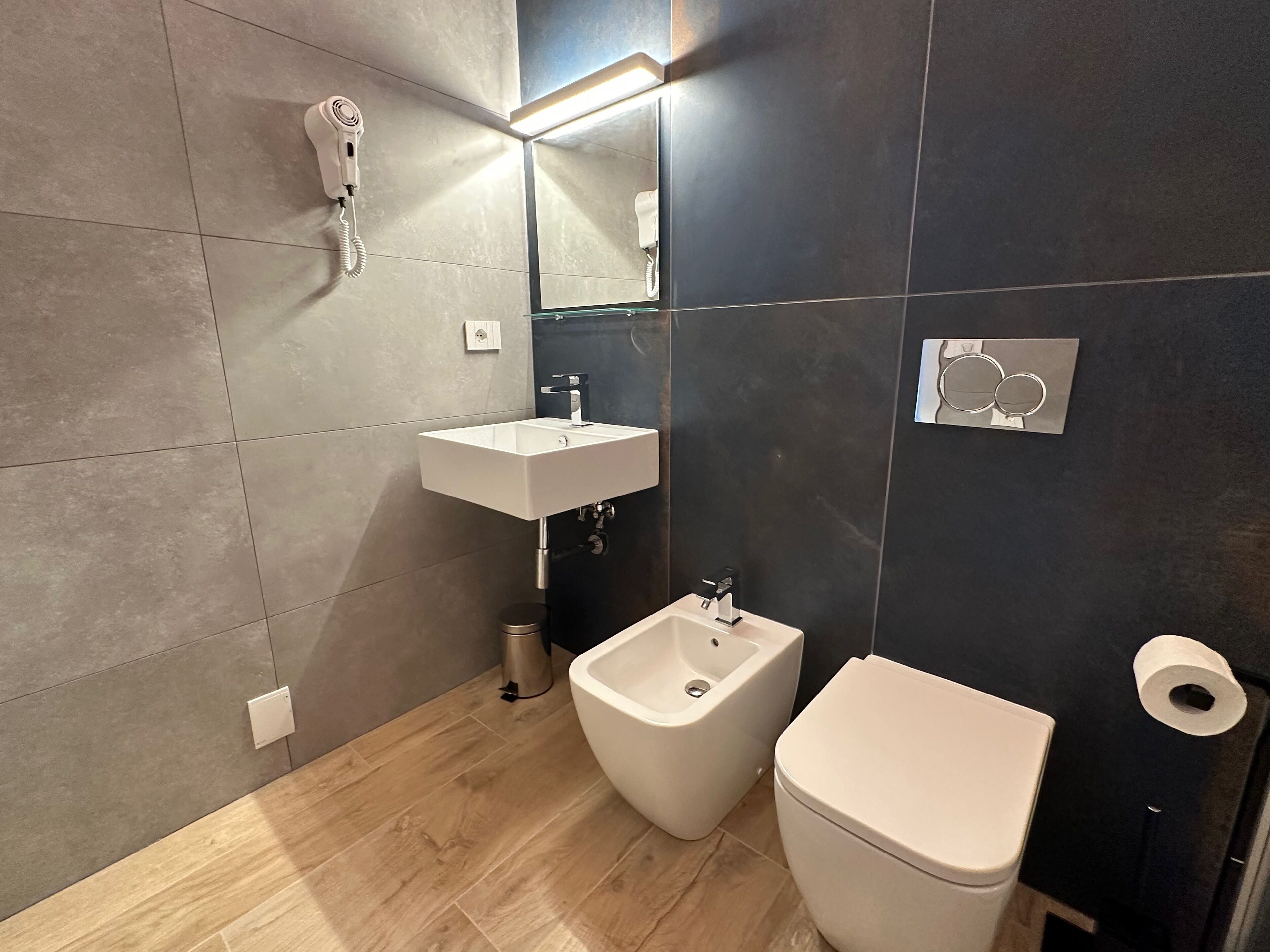 Et moderne badeværelse med hvid vask, bidet og toilet, en hårtørrer og et spejl.