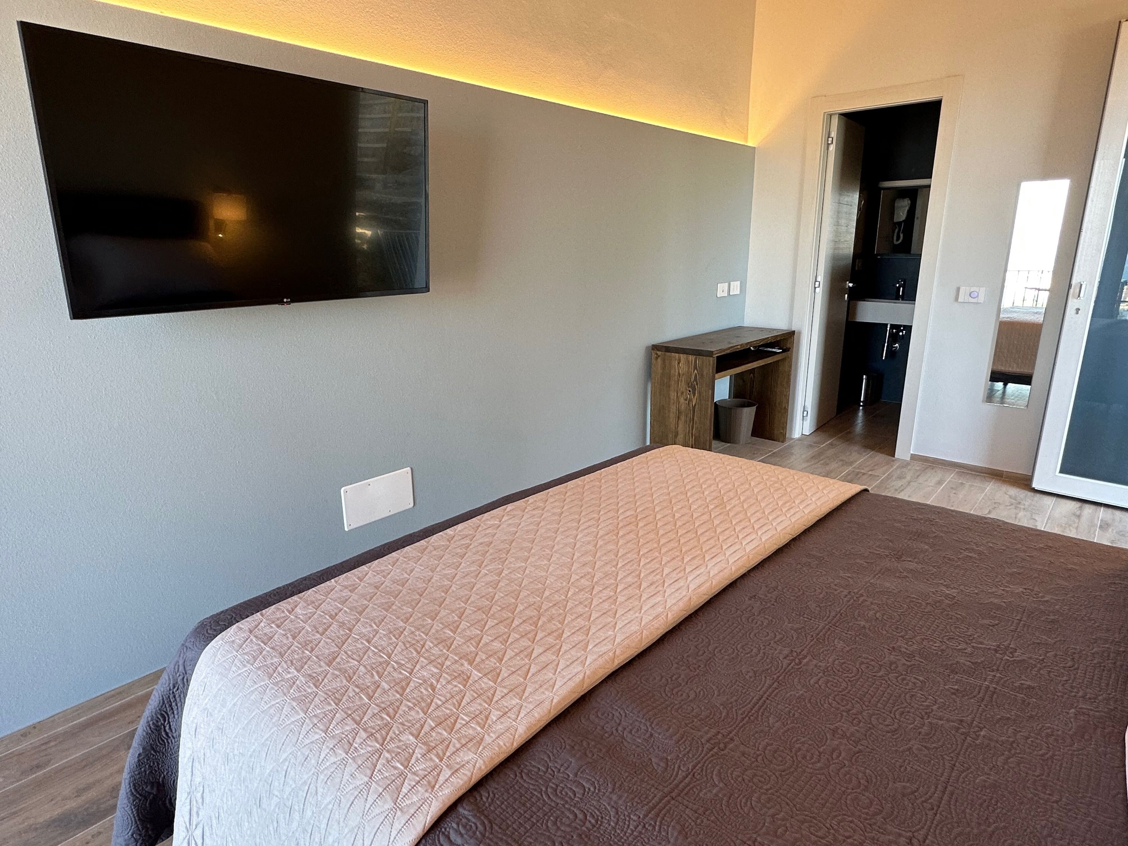 A modern hotel room with a large bed, a flat-screen TV mounted on the wall, a small wooden table, and a kitchenette in the background.