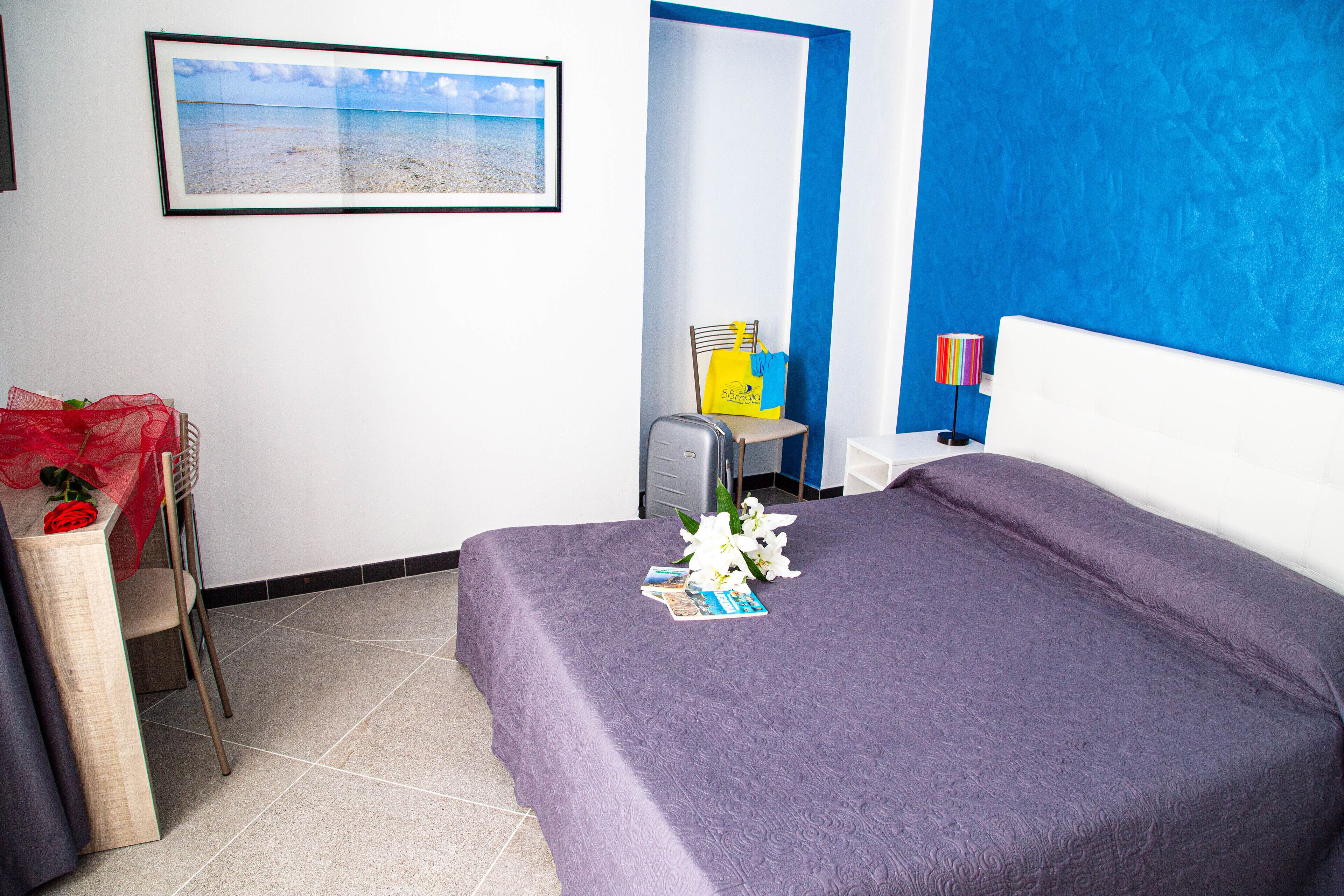 A hotel room with a bed, a chair, a desk, a framed picture on the wall, and a blue accent wall.