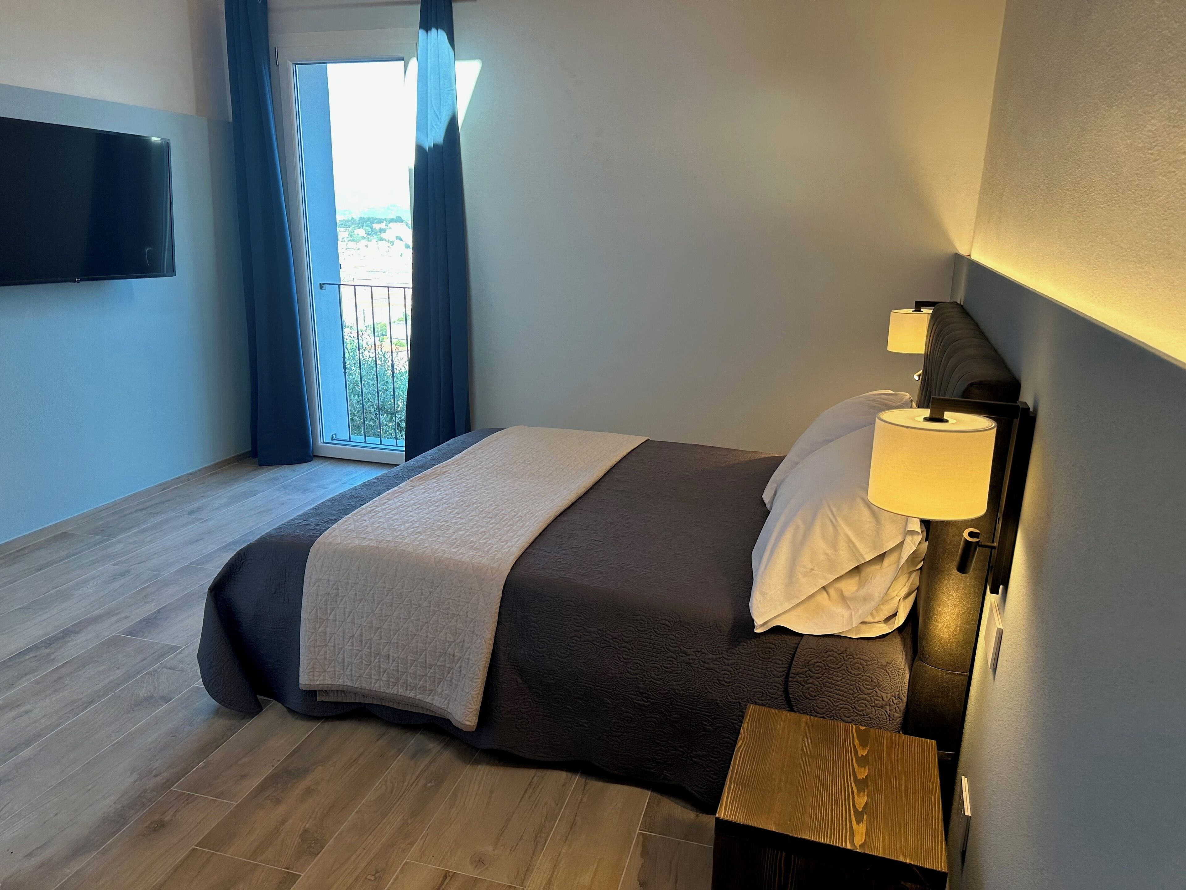 Habitación de hotel con cama, mesita de noche, lámpara, televisor montado en la pared y vista al exterior a través de una ventana.