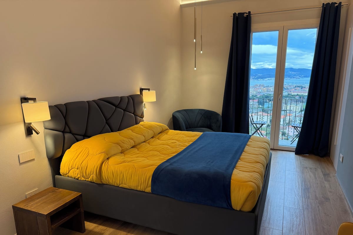 Une chambre moderne avec un grand lit, une table de chevet, une chaise et une vue sur le paysage urbain depuis un balcon.