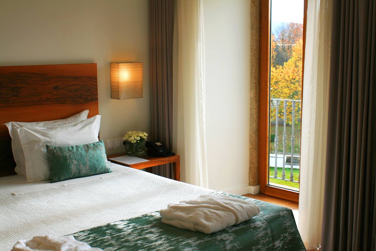 Une chambre d’hôtel avec un grand lit, une table de chevet avec une lampe et une vue sur les arbres aux couleurs automnales par la fenêtre.