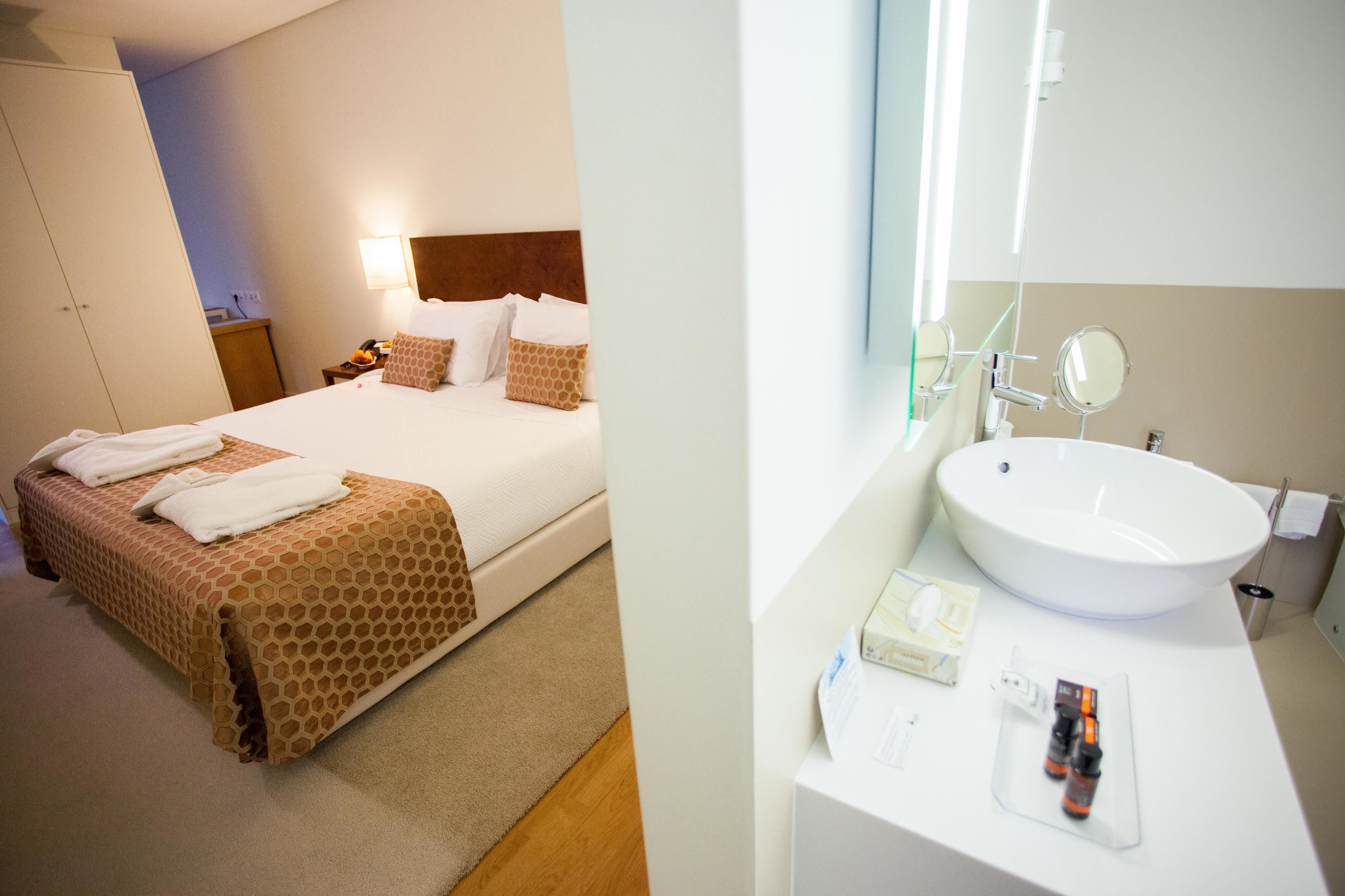 Ein Hotelzimmer mit einem ordentlich bezogenen Bett, einem Nachttisch mit Lampe und einem geräumigen Badezimmerwaschbecken mit Pflegeprodukten.