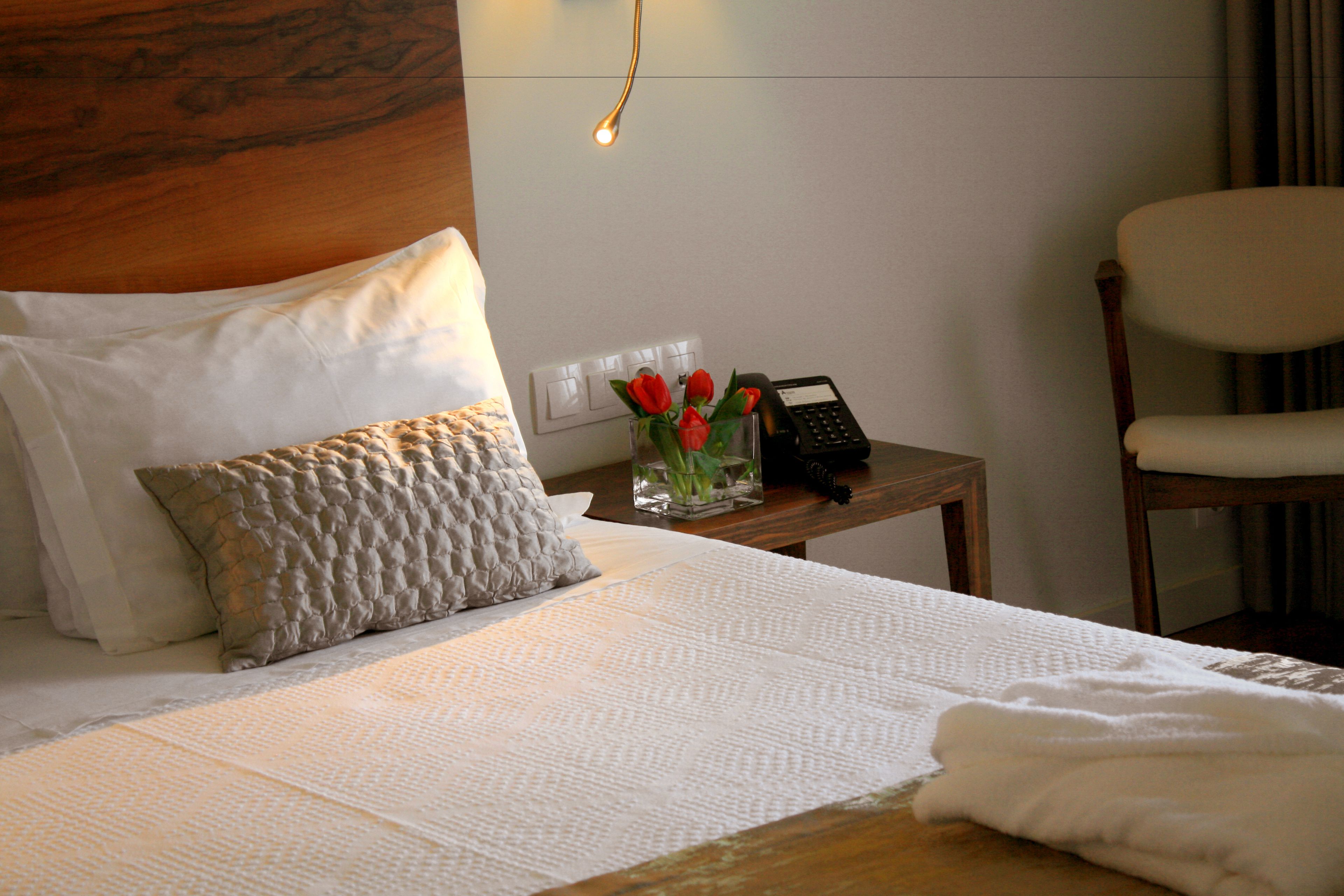 A neatly made bed with white linens, a wooden headboard, and a bedside table with a vase of red roses and a telephone.