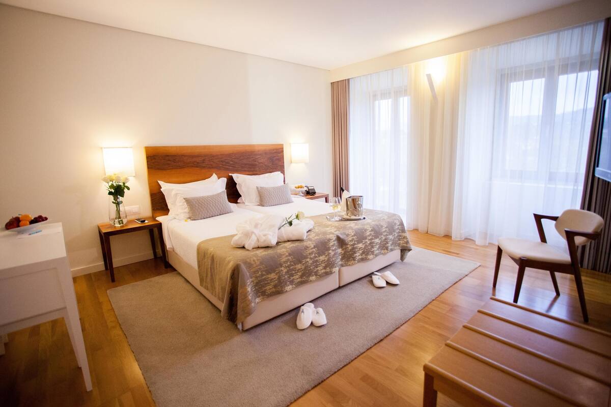 Une chambre d’hôtel avec un grand lit, une table de chevet avec une lampe, une chaise et une vue sur l’extérieur par la fenêtre.