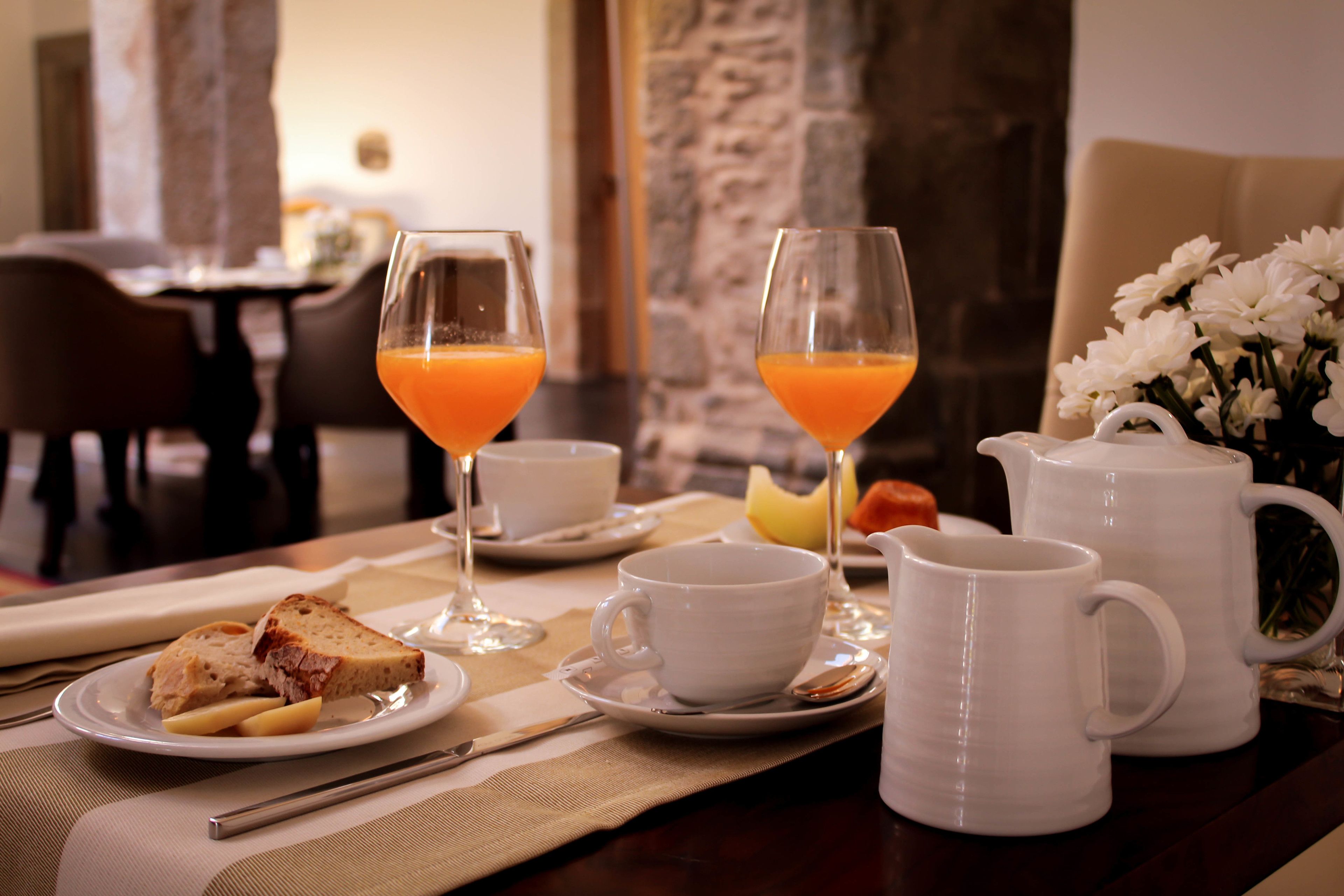 A breakfast setting with a plate of toast, two glasses of orange juice, a white mug, a white pitcher, and a vase of white flowers on a table.