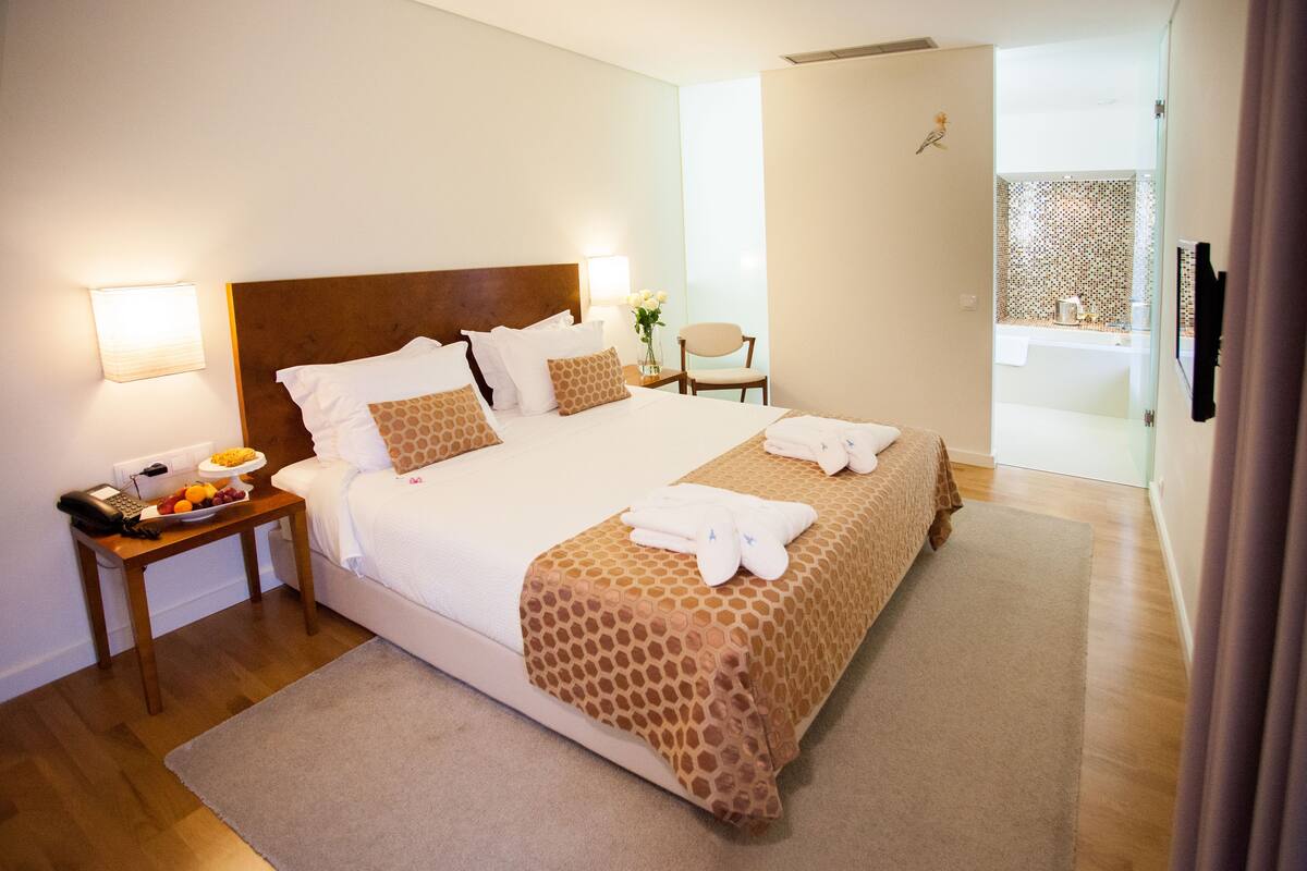 Une chambre d’hôtel avec un grand lit, une table de chevet sur laquelle se trouve un panier de fruits, une chaise et une salle de bain visible à travers une porte ouverte.