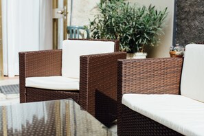Mobiliario de mimbre con cojines blancos, una mesa de cristal y una planta en maceta al fondo.