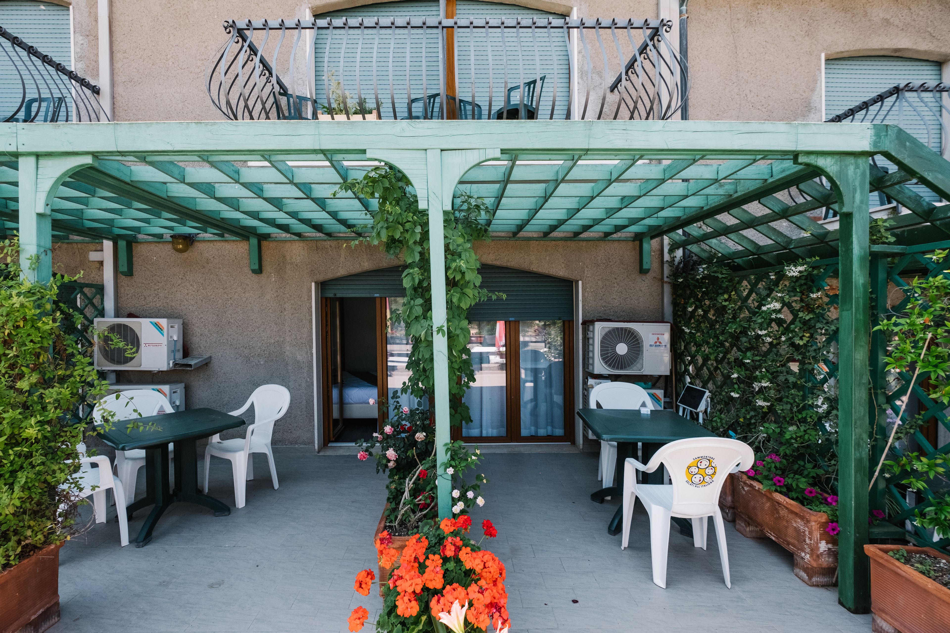 Patio esterno con tavoli e sedie, circondato da vasi con piante e fiori.