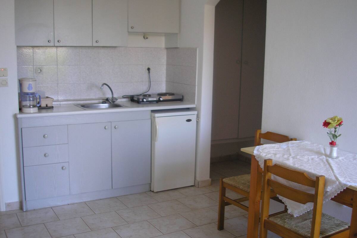A compact kitchen with white cabinets, a sink, a small refrigerator, and a dining table with a white tablecloth.