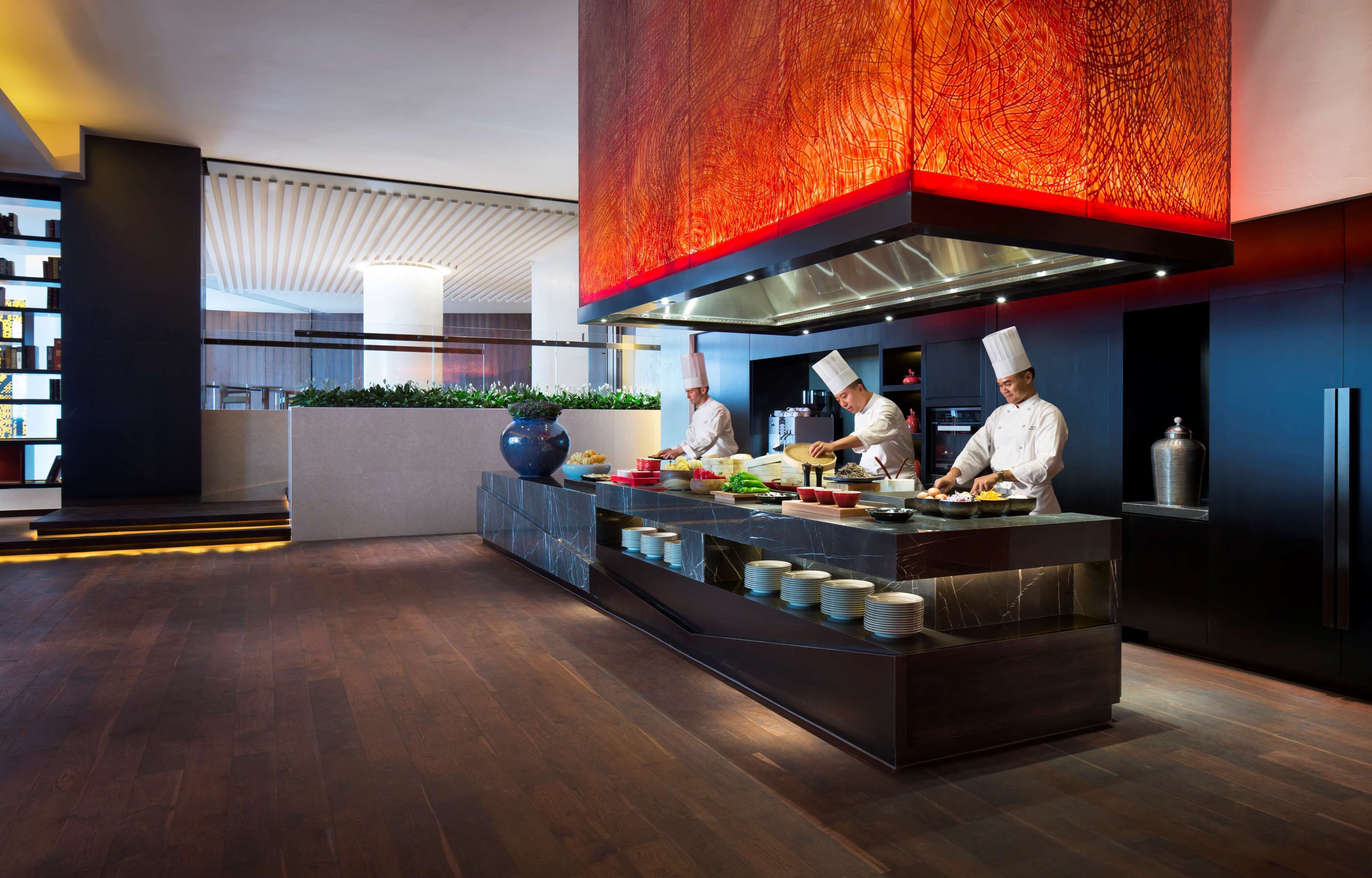 A modern kitchen with chefs preparing food, a marble countertop, and a red accent wall.