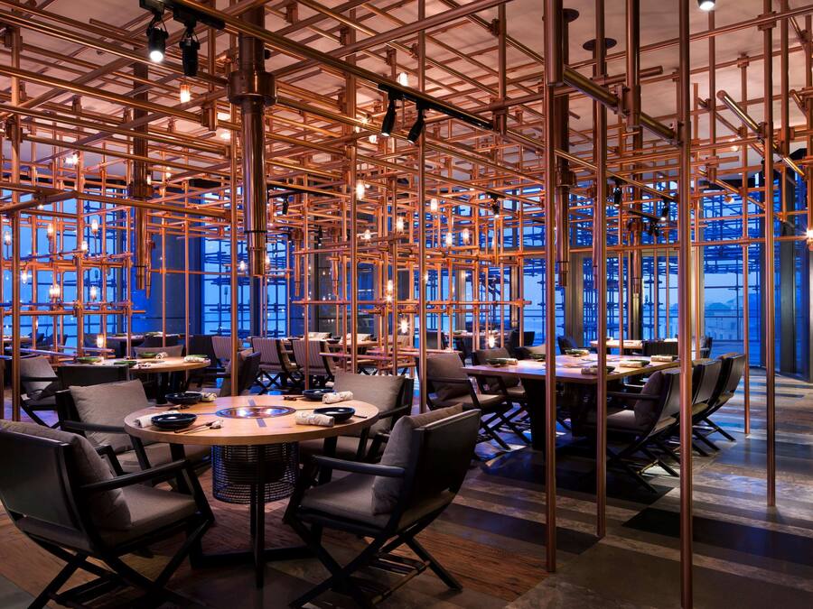 A modern restaurant with a high ceiling and exposed copper beams, featuring round tables and black leather chairs.