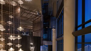 Un moderno vestíbulo de hotel con techo alto, candelabros de cristal colgantes, una escalera central y vistas de un puente de noche.