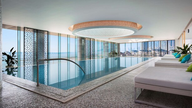 Un área de piscina interior moderna con barandillas de vidrio, vista despejada del paisaje urbano y una fila de sillones con cojines.