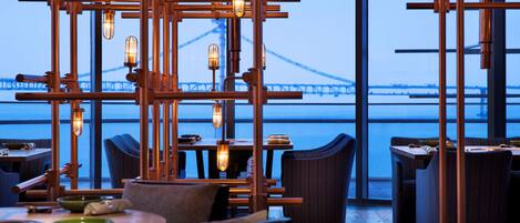 A restaurant with a view of a bridge, featuring exposed copper fixtures and wicker furniture.