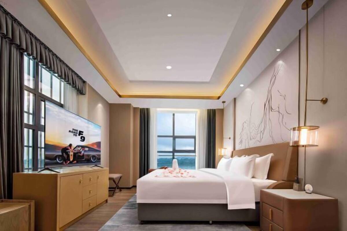 Ein modernes Hotelzimmer mit einem großen Bett, einem Fernseher auf einer Holzkommode, Blick aufs Meer und einer Wand mit einer einfachen Linienzeichnung.