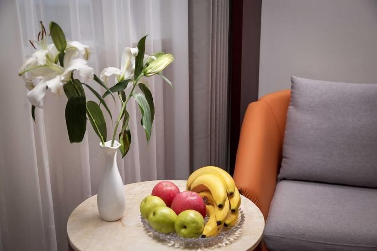 Una mesa redonda con un jarrón blanco con flores blancas, un frutero con manzanas y plátanos, y un cojín gris en una silla naranja.