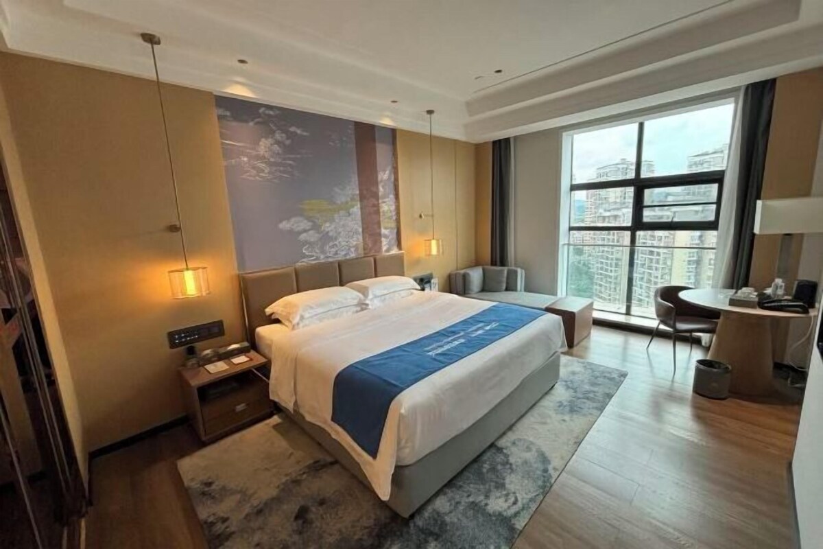 A hotel room with a large bed, a desk, a chair, and a window with a city view.