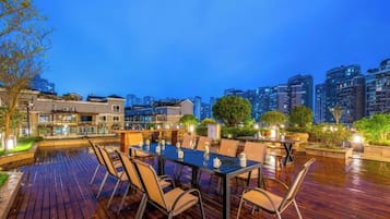 Un espacio al aire libre con mesa y sillas, rodeado de plantas y vistas a la ciudad al anochecer.
