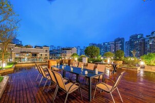 Un espacio al aire libre con mesa y sillas, rodeado de plantas y vistas a la ciudad al anochecer.