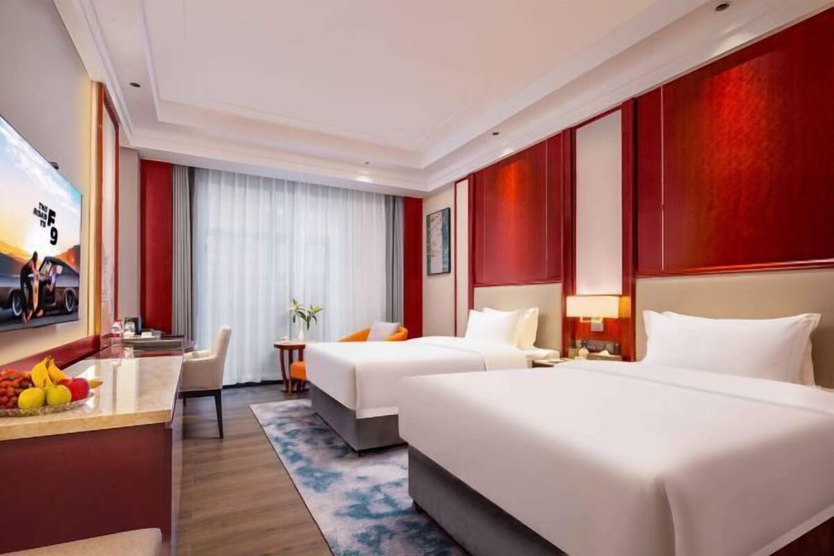 Ein Hotelzimmer mit zwei Betten, einem Schreibtisch mit Früchten, einem Fernseher und einem Fenster mit Vorhängen.
