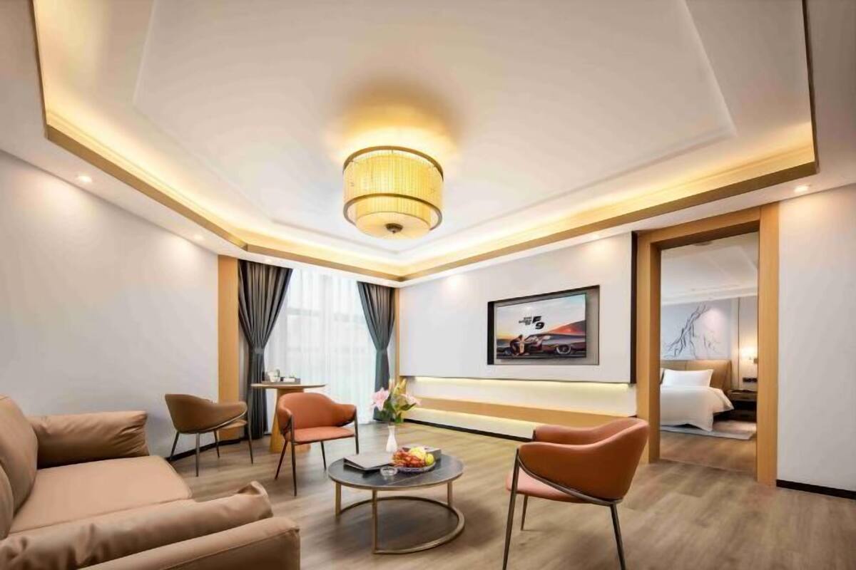 Ein modernes Hotelzimmer mit einer Couch, zwei Sesseln, einem Couchtisch mit Früchten, einem Bild an der Wand und einem Bett mit einem Nachttisch.