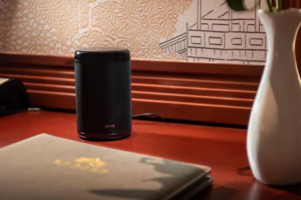 Un altavoz negro sobre una mesa de madera, con un jarrón blanco y un papel tapiz estampado al fondo.