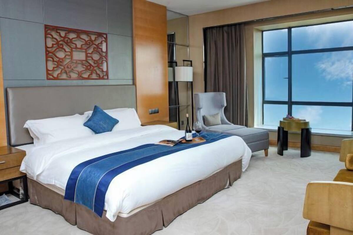 A hotel room with a large bed, a chair, a lamp, and a view of the sky through a large window.