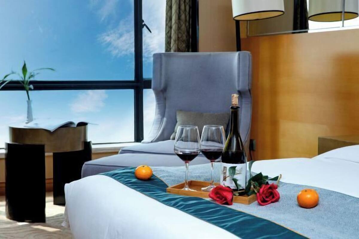 A hotel room with a bed, a large window, a chair, a table with wine glasses, a bottle, and two oranges.