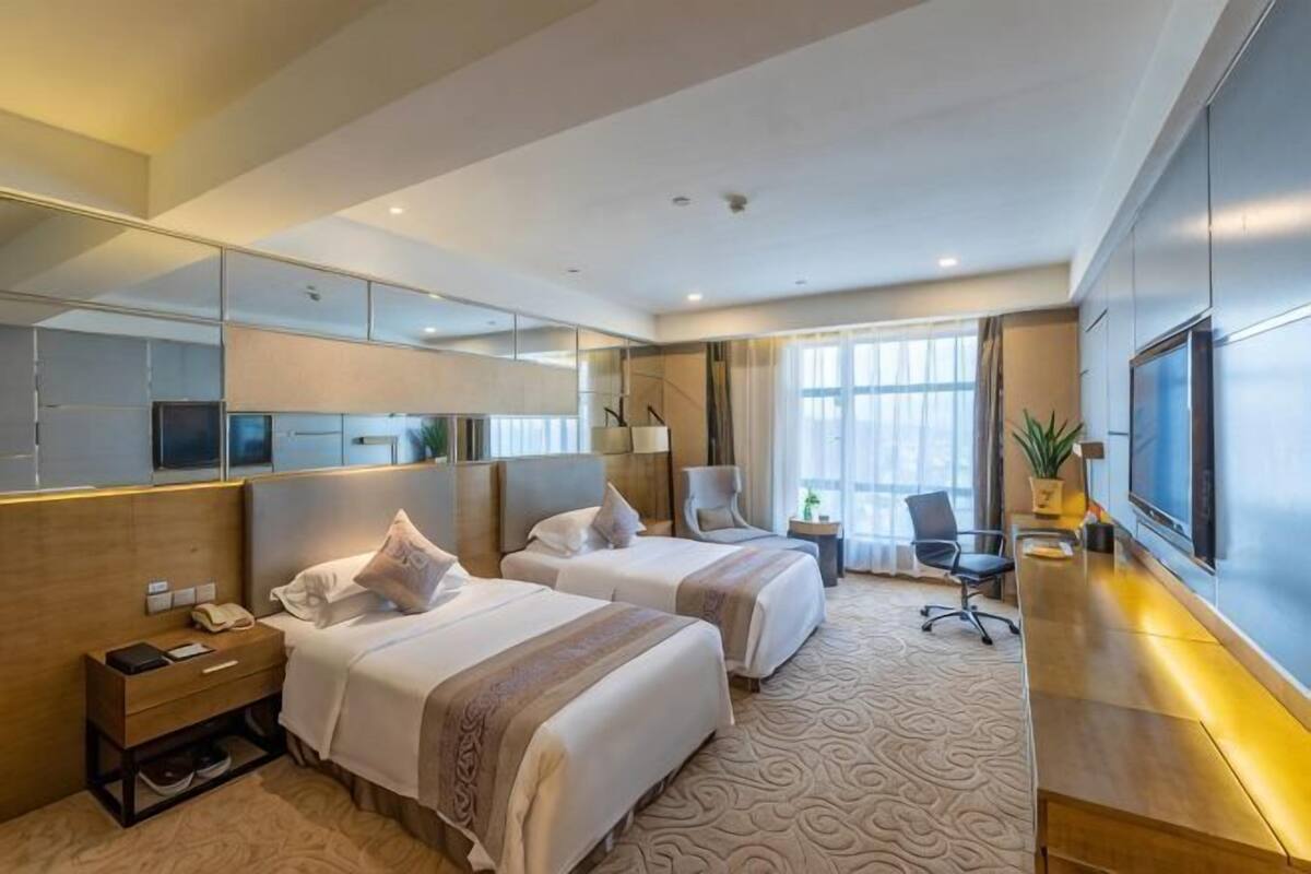 A hotel room with two beds, a desk, a chair, a TV, and a large window.