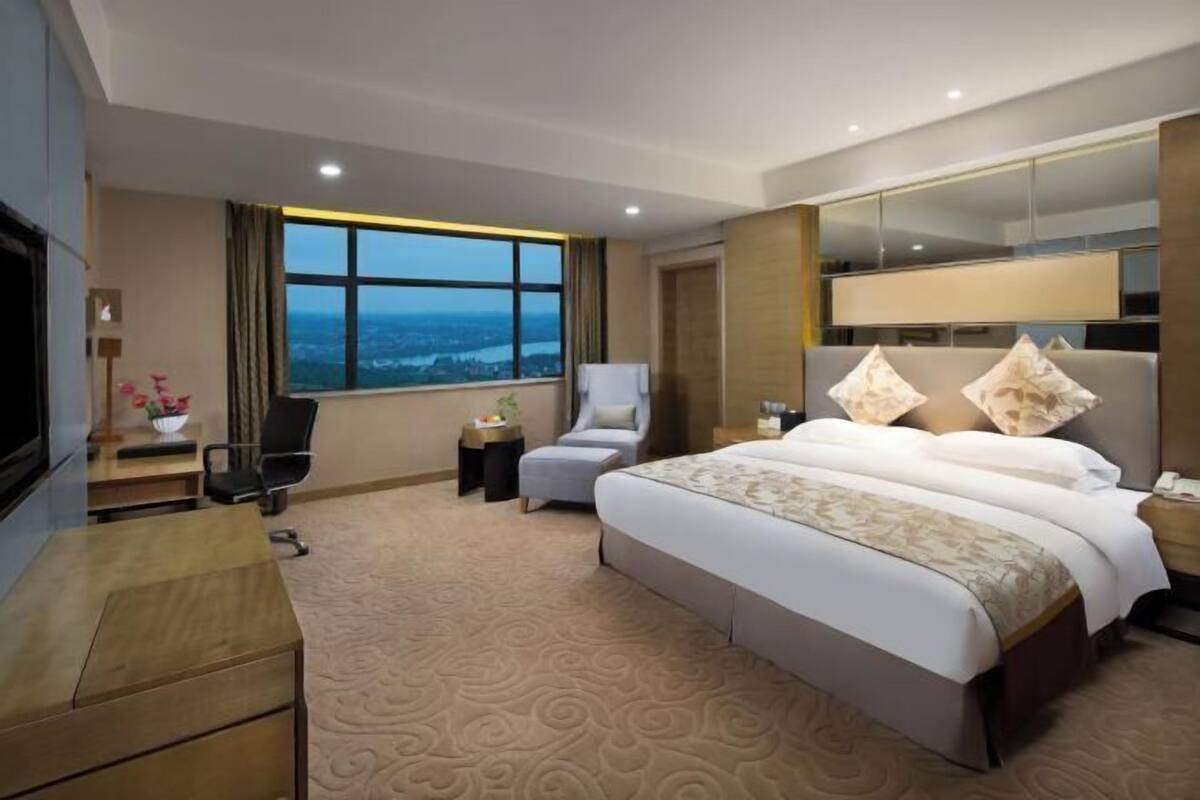 A hotel room with a large bed, a desk, a chair, a small table, a mirror, and a window with a view.